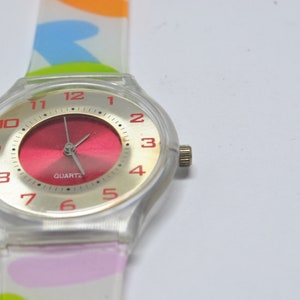 May include: A white plastic watch with a red face and gold-colored numbers. The watch has a colorful patterned band.