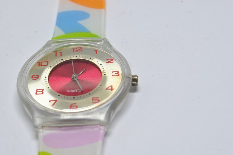 May include: A white plastic watch with a red face and gold-colored numbers. The watch has a colorful patterned band.