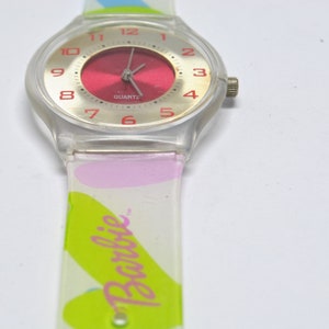 May include: A clear plastic watch with a red face and silver hands. The watch band is white with pink and green accents and the word "Barbie" printed on it.