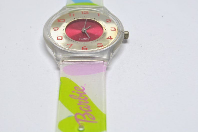 May include: A clear plastic watch with a red face and silver hands. The watch band is white with pink and green accents and the word "Barbie" printed on it.