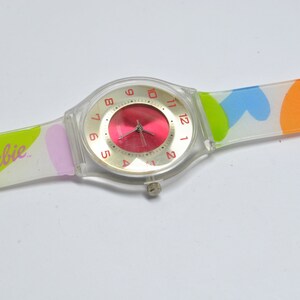 May include: A clear plastic watch with a red face and a multi-colored band. The band has a heart pattern with pink, blue, green, and orange colors. The watch face has the word "Barbie" printed on it.