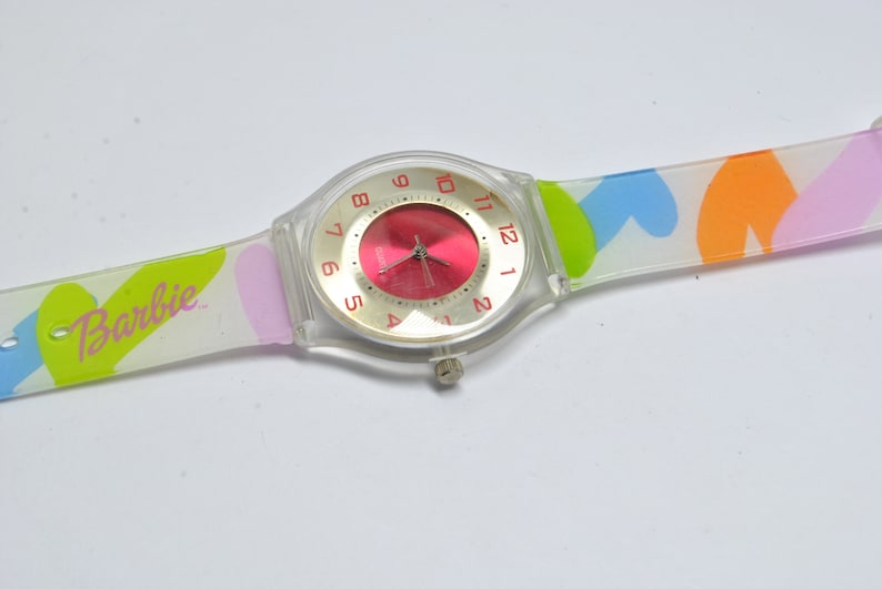 May include: A clear plastic watch with a red face and a multi-colored band. The band has a heart pattern with pink, blue, green, and orange colors. The watch face has the word "Barbie" printed on it.