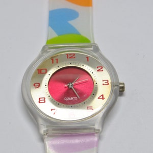May include: A clear plastic wristwatch with a red face and gold-colored numbers. The watch has a colorful, patterned band.