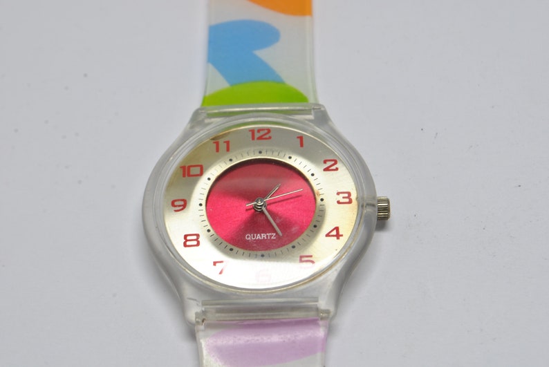 May include: A clear plastic wristwatch with a red face and gold-colored numbers. The watch has a colorful, patterned band.