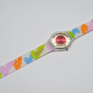 May include: A clear plastic watch with a pink face and a white band with a colorful heart pattern. The band has the word "Barbie" printed on it.