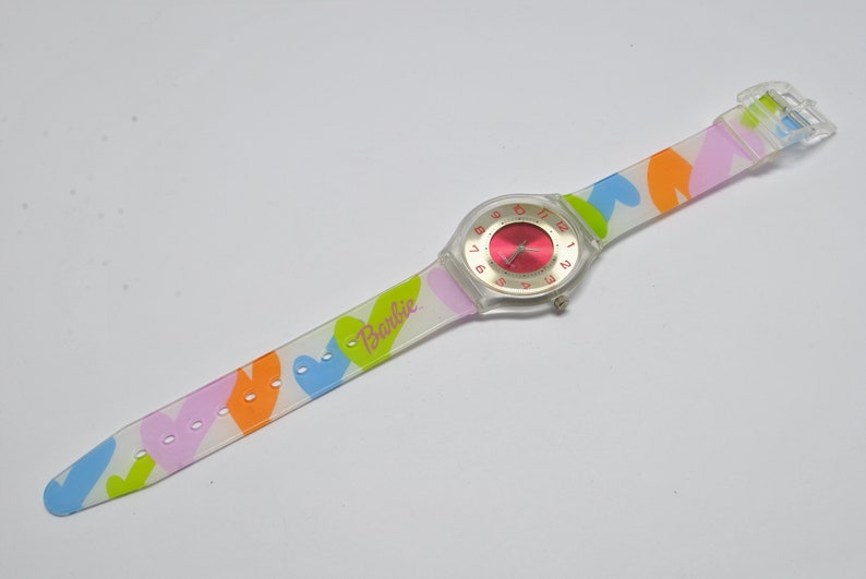 May include: A clear plastic watch with a pink face and a white band with a colorful heart pattern. The band has the word "Barbie" printed on it.