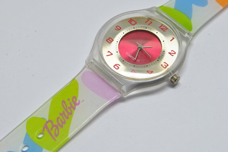 May include: A clear plastic watch with a pink face and white numbers. The watch band is white with a colorful heart pattern and the word "Barbie" printed on it.