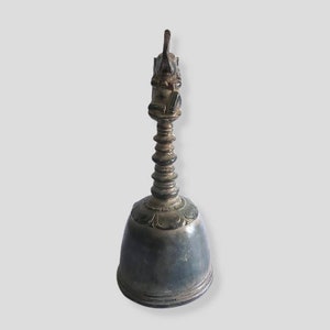 Può includere: Una campana vintage in bronzo con un manico dettagliato con una figura decorativa. La campana ha una base svasata con dettagli ornati e uno stelo scanalato. La campana è di un colore bronzo tenue.