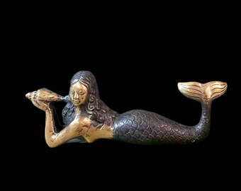 May include: A bronze mermaid figurine, holding a seashell. The mermaid has a gold-colored face and upper body, with a dark, scaled tail. The tail is tipped with gold. The figurine is set against a black background.