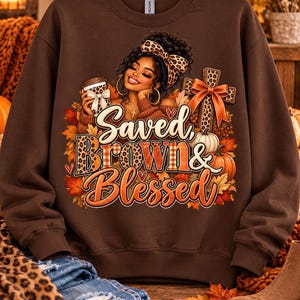 May include: A dark brown crewneck sweatshirt featuring a graphic print of a person with a leopard print headband holding a coffee cup, a cross with a bow, pumpkins, and autumn leaves. The text reads Saved, Brown & Blessed in stylized lettering.