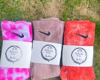 May include: Three pairs of tie-dye socks with a Nike swoosh logo. The socks are pink, brown, and orange. Each pair is packaged with a white label that says "The Rainbow the dye By Ame."
