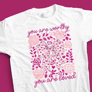 May include: White t-shirt with a pink bow and floral design. The text "you are worthy" and "you are loved" is printed on the shirt.