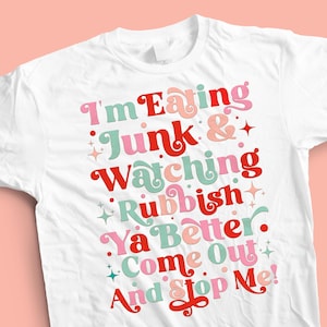 May include: White t-shirt with a colorful graphic design that says "I'm Eating Junk & Watching Rubbish Ya Better Come Out And Stop Me!"