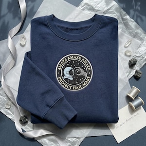 May include: Navy blue sweatshirt with a circular patch featuring an astronaut helmet and a hand reaching out. The patch reads "AMAZE AMAZE AMAZE" and "PROJECT HAIL MARY". The sweatshirt is folded on a textured surface.