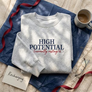 Può includere: Una felpa grigio chiaro con la scritta "HIGH POTENTIAL (currently resting)" in blu navy e rosso. La felpa è su una superficie blu con una tazza di caffè, occhiali e un biglietto con la scritta "Recharging...".