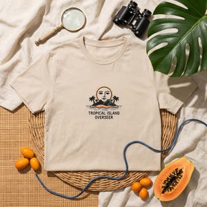 May include: Beige t-shirt with a graphic of an island with a face, palm trees, and the text "TROPICAL ISLAND OVERSEER." The shirt is surrounded by tropical fruits, binoculars, and a magnifying glass.