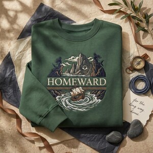May include: Green sweatshirt with an embroidered design featuring a ship sailing through swirling water, mountains, and the word "HOMEWARD". The design includes an eye and figures on the cliffs. The sweatshirt has a crew neck and long sleeves.