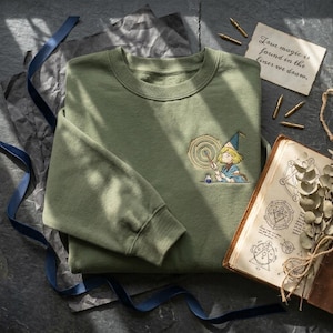 Può includere: Felpa verde oliva con un ricamo di una ragazza maga. Il design include un mago con una bacchetta, un cerchio vorticoso e il testo "True magic is found in the lines we draw" su una piccola carta. Sono visibili anche un libro vintage e penne calligrafiche.