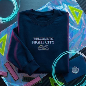 May include: Navy blue sweatshirt with the text "WELCOME TO NIGHT CITY" and a neon motorcycle graphic. The sweatshirt is folded and surrounded by neon green and blue accents, along with computer components and an envelope.