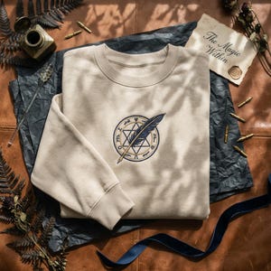 May include: Beige crewneck sweatshirt with an embroidered design of a quill and a six-pointed star inside a circle. The text "The Magic Within" is on a small card. The sweatshirt is on a brown leather surface.