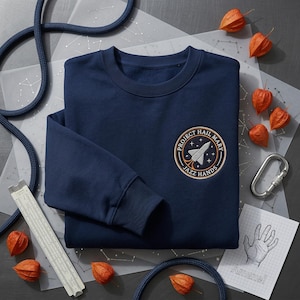May include: Navy blue sweatshirt with a circular patch featuring a rocket ship and the words "PROJECT HAIL MARY" and "JAZZ HANDS." The sweatshirt is surrounded by orange dried flowers, a slide rule, and a drawing that says "Amaze!"