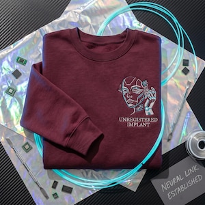 May include: A maroon crewneck sweatshirt with a futuristic design. The design features a stylized face with neon blue accents and the text "UNREGISTERED IMPLANT". The words "NEURAL LINK ESTABLISHED" are on a small gray card.