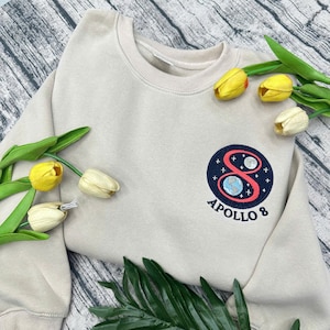May include: A beige crew neck sweatshirt with an embroidered Apollo 8 patch. The patch features a red number 8, a blue and white planet, and the words "APOLLO 8" in white. Yellow tulips and green leaves are arranged around the sweatshirt.