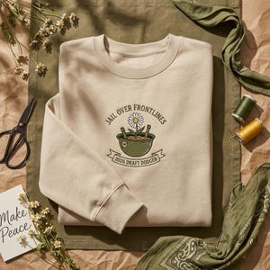 May include: A beige sweatshirt with the text "JAIL OVER FRONTLINES" and "2026 DRAFT DODGER" surrounding a flower in a pot. The sweatshirt is folded on a brown surface with scissors, flowers, and spools of thread.