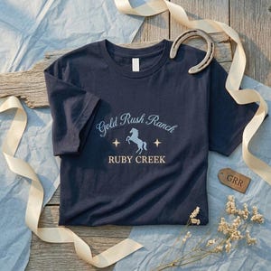 Könnte beinhalten: Marineblaues T-Shirt mit dem Text "Gold Rush Ranch Ruby Creek" und einer Pferde-Grafik. Ein Hufeisen, ein Band und ein kleines Etikett sind um das Shirt angeordnet. Das Shirt ist aus einem weichen Material.