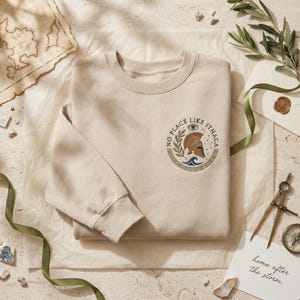 May include: Beige crewneck sweatshirt with a circular graphic featuring a helmet, olive branch, and text that reads "NO PLACE LIKE ITHACA." The design is in shades of blue, brown, and beige. The sweatshirt is folded on a cream-colored surface.