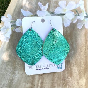 May include: A pair of teardrop-shaped earrings with a metallic teal finish and a textured snakeskin pattern. The earrings are attached to a white display card featuring the text THE TEAL CHEETAH Earrings by Meghan and a small cheetah icon.