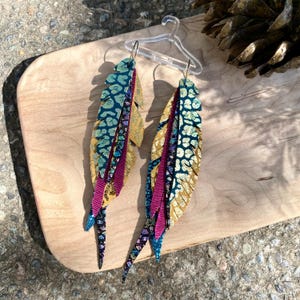 May include: A pair of handmade feather earrings. The earrings feature a layered design with gold, teal, and purple accents. The feathers are attached to a silver hook. The earrings are displayed on a wooden surface.