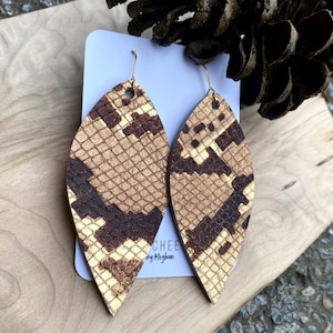 May include: A pair of teardrop-shaped earrings with a snakeskin pattern in shades of brown, tan, and cream. The earrings have gold-colored hooks. The earrings are displayed on a white card with the text "CHEE by Meghan".