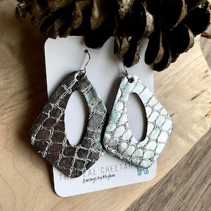 May include: A pair of silver-colored, diamond-shaped earrings with a textured, reptile-like pattern. Each earring has a large, oval cutout in the center and is suspended from a black hook. The earrings are displayed on a white card with text that reads "The Real Cheetah Earrings by Meghan."