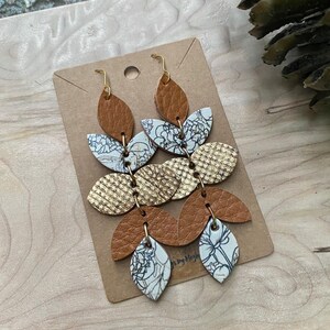 May include: Dangle earrings with a layered leaf design. The earrings feature brown, gold, and floral patterned leather pieces. Gold-colored ear wires and connecting hardware complete the design. The earrings are displayed on a brown card.