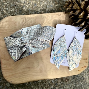 May include: A silver-colored cuff bracelet and a pair of leaf-shaped earrings. The jewelry has a scale-like texture with iridescent highlights. The earrings are displayed on a white card. The items are on a wooden surface.
