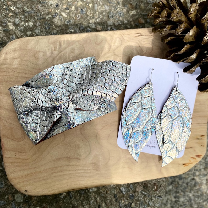 K&ouml;nnte beinhalten: Ein silbernes Armband und ein Paar ohrf&ouml;rmige Ohrringe. Der Schmuck hat eine schuppenartige Textur mit schillernden Highlights. Die Ohrringe werden auf einer wei&szlig;en Karte pr&auml;sentiert. Die Artikel befinden sich auf einer Holzoberfl&auml;che.