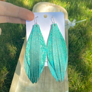 May include: A pair of long, teardrop-shaped earrings featuring a vibrant turquoise metallic finish with a subtle snakeskin-like texture. The earrings are constructed from layered, thin vertical strips of material, creating a fringe effect.