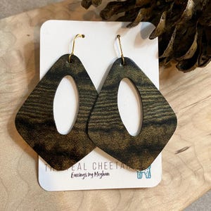 May include: A pair of diamond-shaped earrings with a gold and black pattern, featuring an oval cutout in the center. The earrings hang from gold-colored hooks. The text on the card reads "THE REAL CHEETAH Earrings by Meghan."