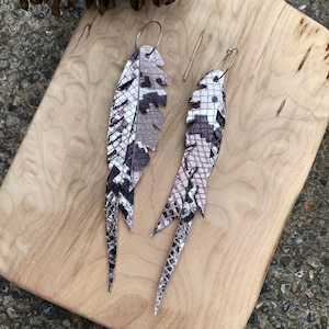 May include: A pair of feather-shaped earrings with a snakeskin pattern in shades of gray, white, and black. The earrings are made of a textured material and have silver-colored hoops. The earrings are displayed on a light-colored wooden surface.