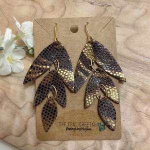 May include: A pair of gold and brown leather earrings with a leaf design. The earrings are made by The Teal Cheetah Earrings by Meghan.