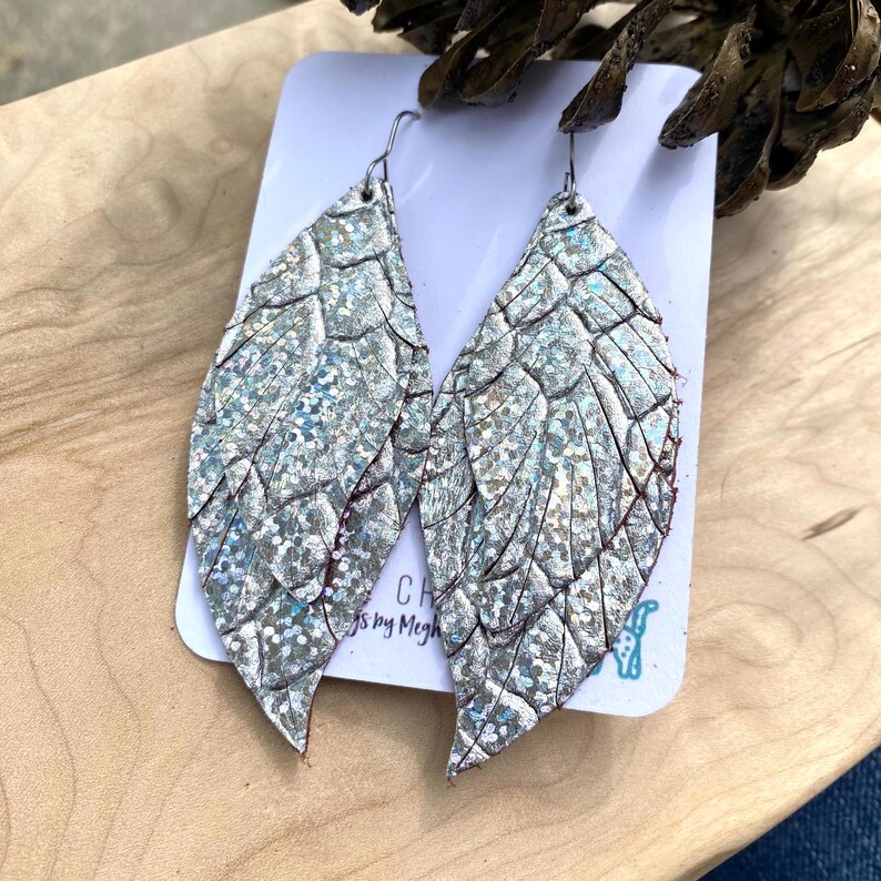K&ouml;nnte beinhalten: Ein Paar silberne, blattf&ouml;rmige Ohrringe mit strukturierter, glitzernder Oberfl&auml;che. Die Ohrringe h&auml;ngen an einer wei&szlig;en Karte mit dem Text "CH by Megh". Die Ohrringe haben silberne Haken.
