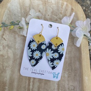 May include: A pair of teardrop-shaped earrings featuring a black background with a white daisy pattern and a gold semi-circular top piece. The earrings are attached to a white display card labeled THE TEAL CHEETAH Earrings by Meghan.