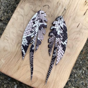 May include: A pair of feather-shaped earrings with a snakeskin pattern in black, white, and pale pink. The earrings are made of leather and have silver-colored ear wires. The earrings are displayed on a wooden surface.