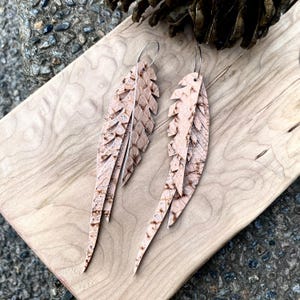 May include: A pair of feather-shaped earrings. The earrings are made of light brown leather with a textured pattern and silver-colored ear wires. The earrings are displayed on a light wooden surface.