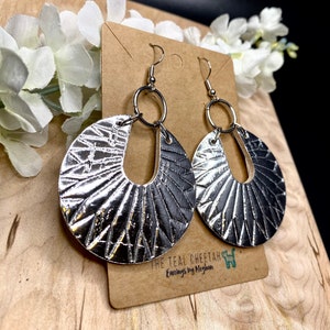 May include: Silver tone earrings with a textured, geometric design. The earrings are in the shape of a circle with a cut-out in the center. The earrings are hanging from silver hooks. The earrings are displayed on a brown card with the text "The Teal Cheetah Earrings by Meghan".