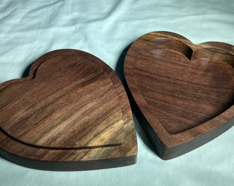 Puede incluir: Dos cajas de madera en forma de corazón, una abierta y otra cerrada, que muestran una veta de madera marrón oscura. Las cajas tienen un interior tallado, ideal para pequeños recuerdos o joyas.