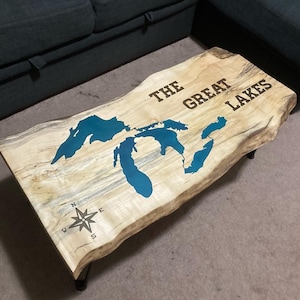 Puede incluir: Una mesa de café de madera con un mapa azul de los Grandes Lagos y el texto "THE GREAT LAKES" en la parte superior. También hay una brújula en la mesa.