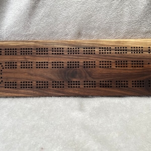 Peut inclure: Un plateau de jeu de cribbage en bois avec une finition en noyer brun foncé. Le plateau a un design traditionnel avec plusieurs rangées de trous pour les pions.