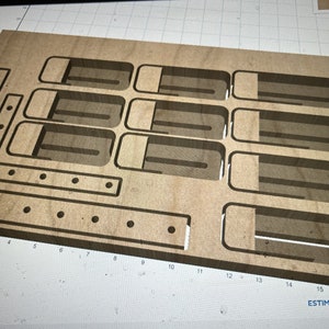 Puede incluir: Un organizador de madera con múltiples compartimentos para guardar objetos pequeños. El organizador tiene forma rectangular y está hecho de madera marrón claro. Tiene un patrón de cuadrícula en la superficie y está marcado con los números del 1 al 16. El texto "ESTIMATE Roughing: 21-25" es visible en la esquina inferior derecha.